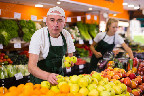 Portrait of cheerful male seller in uniform holding fresh golden apples in grocery shop