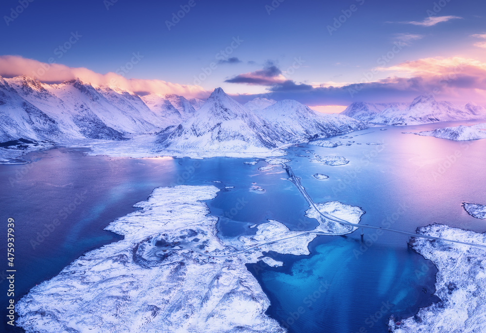 Aerial view of sea, snowy islands, mountains, road, violet sky at sunset in winter. Lofoten islands, Norway. Landscape with mountains and rocks in snow, water at dusk. Top view from drone. Nature