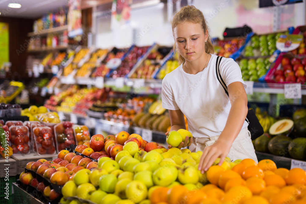Positive girl consumer choosing fresh apples at grocery section of supermarket
