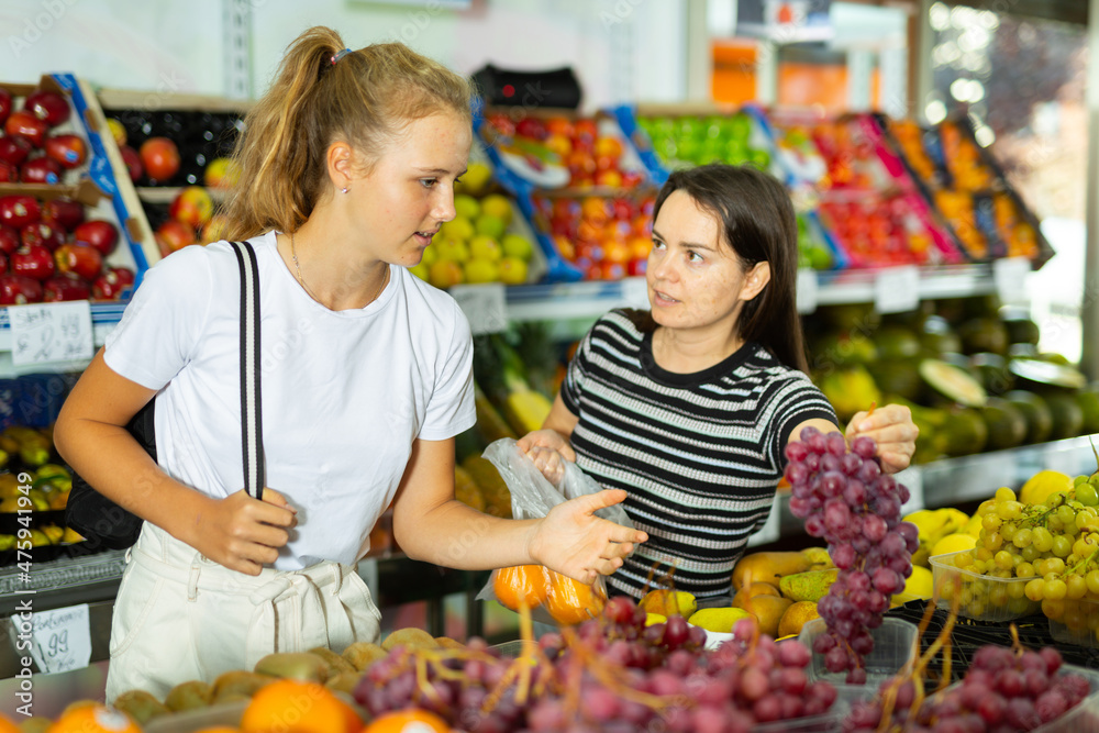 Young woman with a fifteen-year-old girl, who came to the store, attentive choose grapes, standing near the fruit counter
