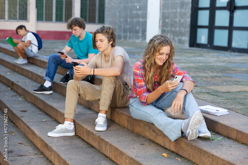 Teenager girl sitting on stairs beside school building and using smartphone. Her classmate sitting next to her and using smartphone too.