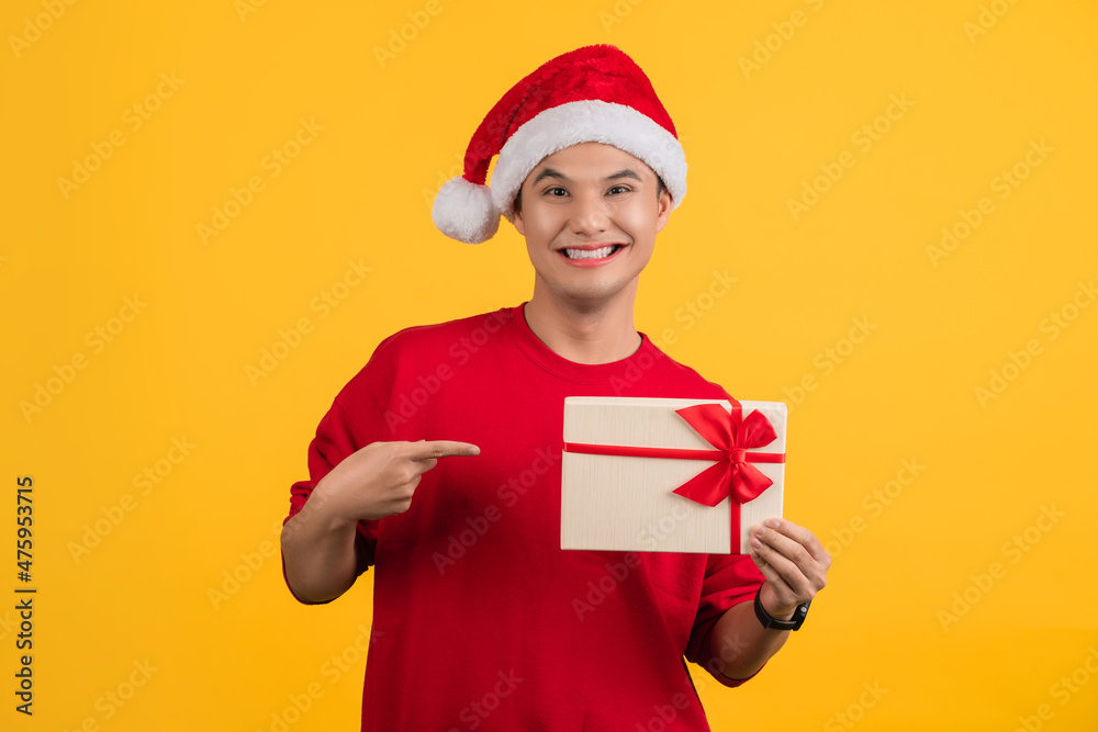 Excited asian handsome man wearing red t-shirt and christmas hat posing isolated on orange background. Mock up copy space. He pointing index finger on gift certificate Excited asian handsome man wearing red t-shirt and christmas hat posing isolated on orange background. Mock up copy space. He pointing index finger on gift certificate