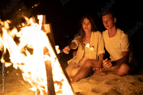 Young couple on the sea beach sitting bonfire and toasting marshmallows on a stick. A romantic date by the fire. Tourism and tourists concept