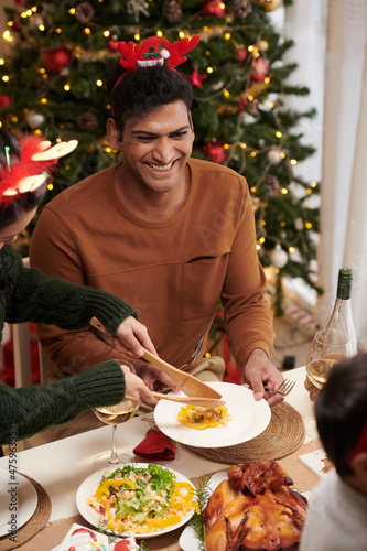 Young man laughing at joke when wife putting salad in his plate at family Christmas dinner