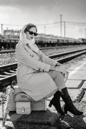 A young woman in a raincoat and dark glasses sits on a suitcase on the platform of a railway station. Retro style black and white outdoor shot