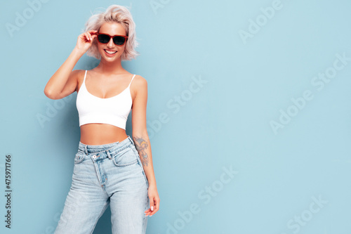 Young beautiful smiling female in trendy summer clothes. Sexy carefree woman posing near blue wall in studio. Positive blond model having fun and going crazy. In sunglasses © halayalex