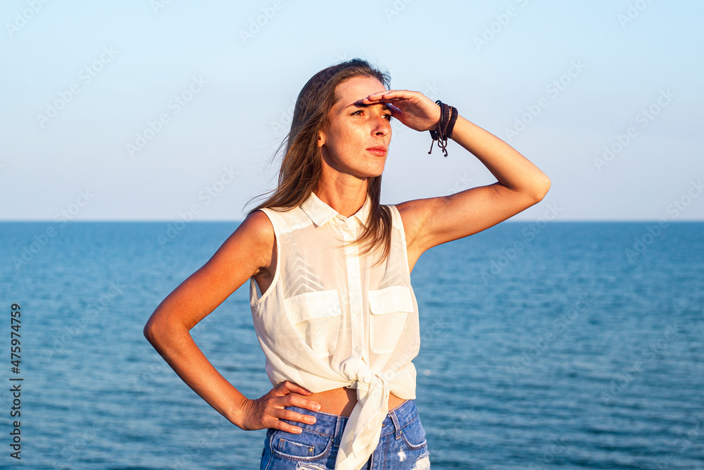 Beautiful young woman putting her palm to her forehead looks into the distance at the sea.