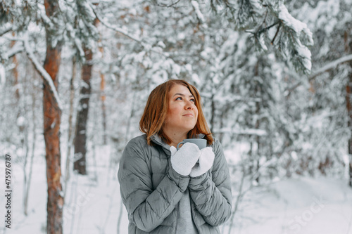 A beautiful girl in a gray jacket and knitted white mittens drinks a drink in a snowy forest, looks up and dreams. Comfort and warmth in the winter season