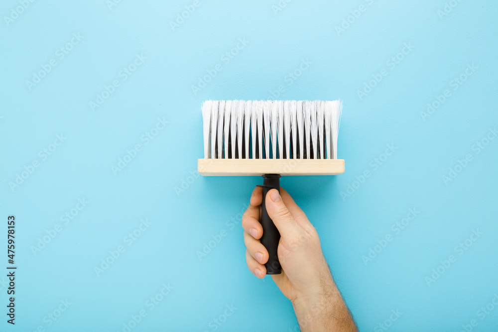 Naklejka premium Young adult man hand holding new brush on light blue table background. Pastel color. Closeup. Preparation for repair work of home. Top down view.