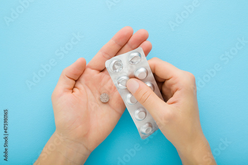 Young adult woman hands holding and opening blister of pills on light blue background. Closeup. Pastel color. Receiving medicaments. Medical, pharmacy and healthcare concept. Top down view.