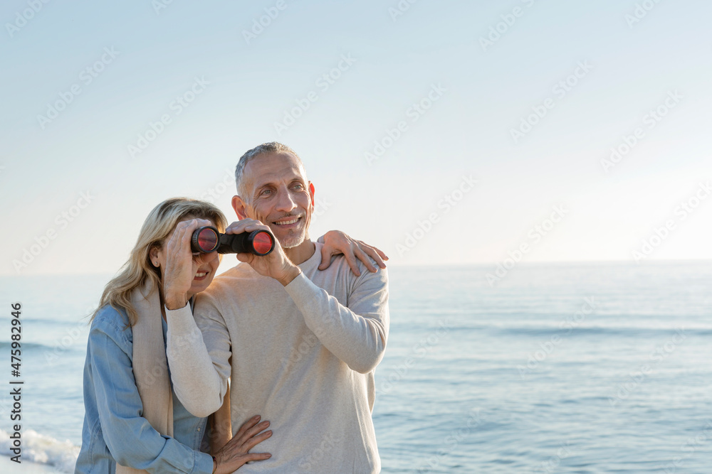 Woman looking through binoculars held by husband at beach Woman looking through binoculars held by husband at beach