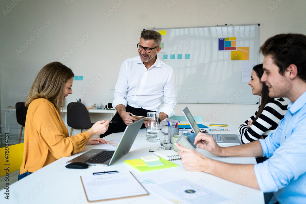 Smiling businessman and colleagues sitting on desk at office Smiling businessman and colleagues sitting on desk at office