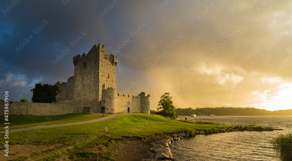 View of the ruins of Ros Castle at sunset under cloudy skies. Medieval castle on the shores of Loch Leane in Killarney National Park, County Kerry.