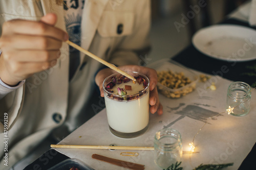 Woman making candle of soy wax, decorating it with dry roses and flowers using wooden stick.