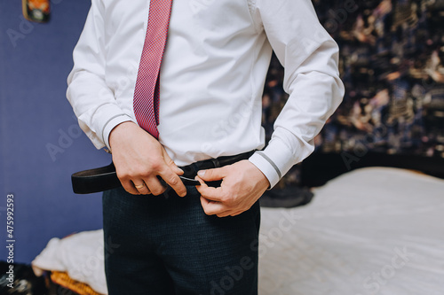 A man, a groom in a white shirt and a red tie puts a leather belt on his trousers, getting ready for a wedding, a wedding in the morning.