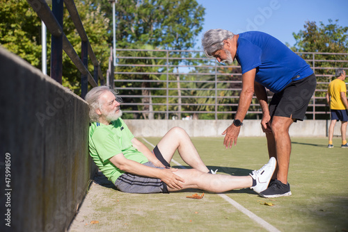 Senior man injured during football training. Caucasian man with grey hair in sport clothes sitting on ground, touching leg, friend helping. Football, sport, leisure concept