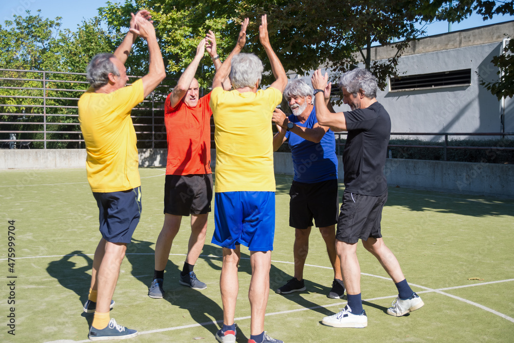Happy senior men having fun on sunny day. Men with grey hair in sport clothes standing in circle on sport field, rising hands. Football, sport, leisure concept