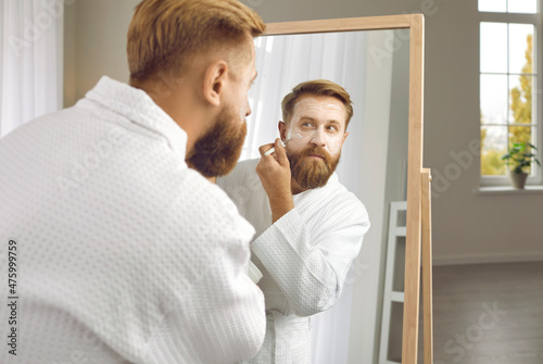 Reflection in mirror of adult man who takes care of himself and applies mask to his face with cosmetic brush. Close up of bearded man in white bathrobe doing beauty treatments in front of large mirror