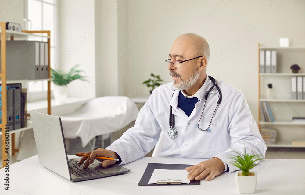 Senior doctor in glasses and white medical gown working on laptop sitting at workplace in office. Serious mature old male doctor uses modern technology to check patient registration forms. Senior doctor in glasses and white medical gown working on laptop sitting at workplace in office. Serious mature old male doctor uses modern technology to check patient registration forms.