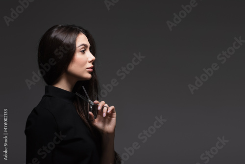 side view of young woman in black turtleneck holding scissors near long shiny hair isolated on dark grey