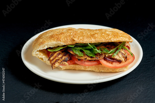 Ciabatta sandwich with pork steak and vegetables in white plate on black background