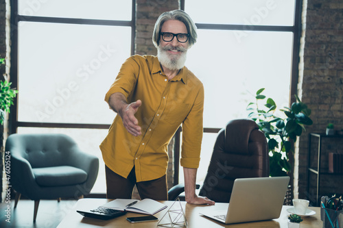 Photo of friendly senior man wear yellow shirt glasses giving you arm for handshake indoors workplace workstation