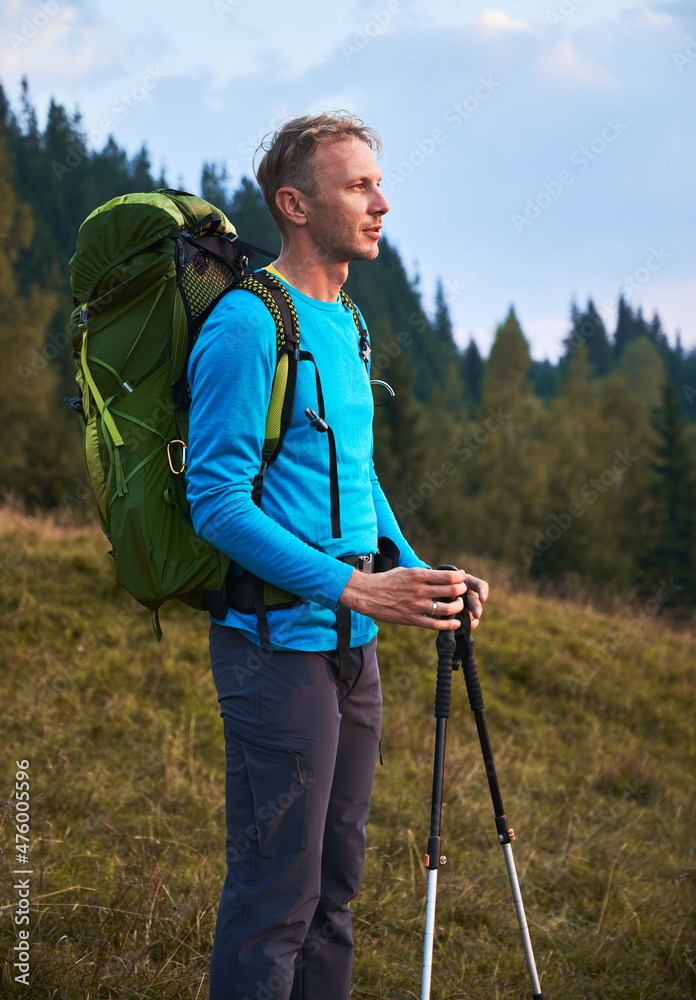 Side view of hiker male with backpack and trekking pole standing on green lawn on the backdrop of forest and watching far away. Sportsman going trek by outdoor spaces. Concept of travelling, hiking. Side view of hiker male with backpack and trekking pole standing on green lawn on the backdrop of forest and watching far away. Sportsman going trek by outdoor spaces. Concept of travelling, hiking.