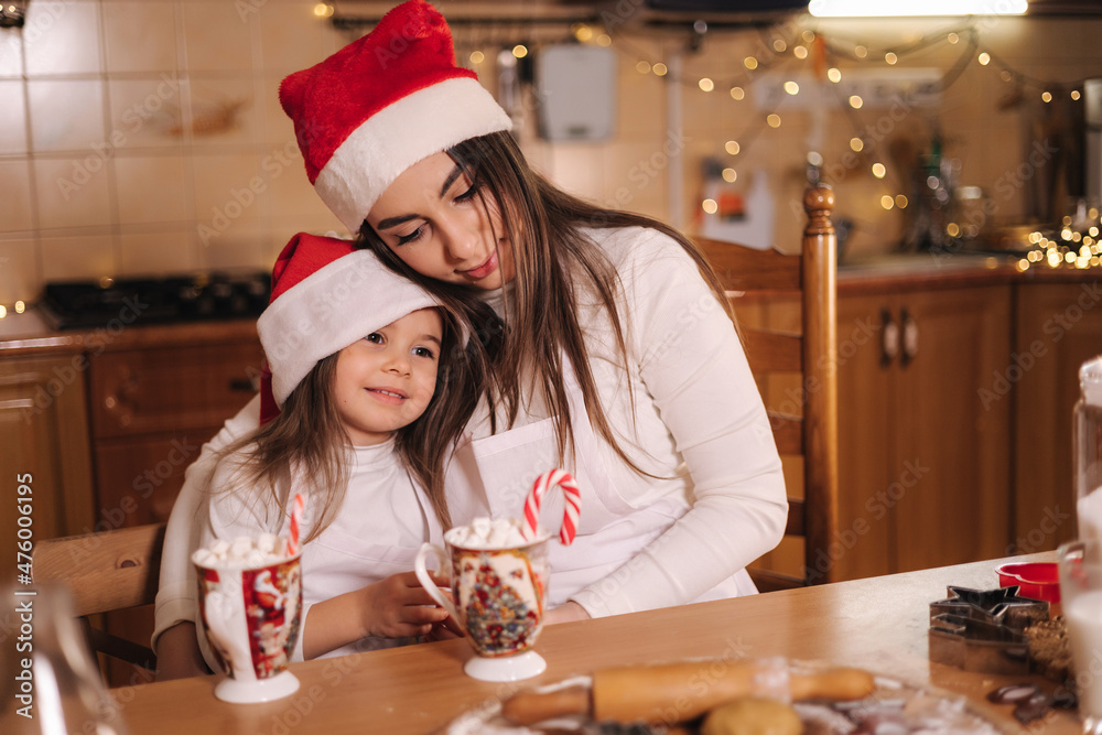 Woman and her little daughter drinking tasty cocoa with marshmallow at the kitchen. Home decorated on Christmas eve. Mom and little girl in santa hat. Background of fairy lights. Fit tree Woman and her little daughter drinking tasty cocoa with marshmallow at the kitchen. Home decorated on Christmas eve. Mom and little girl in santa hat. Background of fairy lights. Fit tree