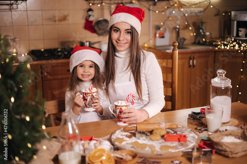 Woman and her little daughter drinking tasty cocoa with marshmallow at the kitchen. Home decorated on Christmas eve. Mom and little girl in santa hat. Background of fairy lights. Fit tree