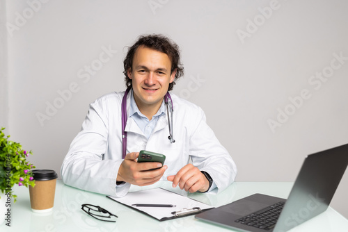 Smiling male doctor using mobile smart phone and working on laptop computer in medical room in the hospital, electronic health records system EHRs, teleconference or telemedicine concept.