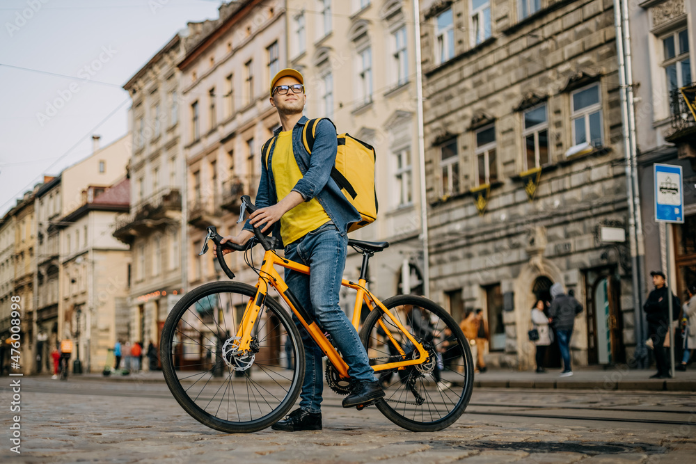 A happy delivery man who is sitting on a bicycle and looking around. He is carrying a yellow backpack on his shoulders. A happy delivery man who is sitting on a bicycle and looking around. He is carrying a yellow backpack on his shoulders.
