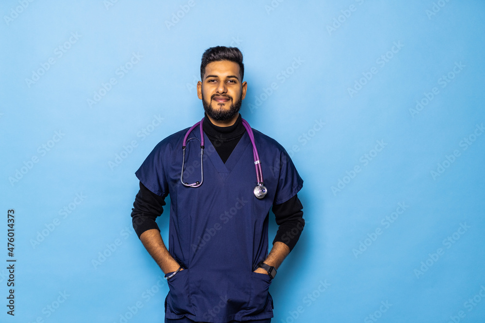 Indian surgeon standing setting his gloves to start the surgery, wearing a mask and a blue uniform, isolated on a blue background. Indian surgeon standing setting his gloves to start the surgery, wearing a mask and a blue uniform, isolated on a blue background.