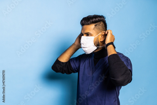 Indian surgeon standing setting his gloves to start the surgery, wearing a mask and a blue uniform, isolated on a blue background.