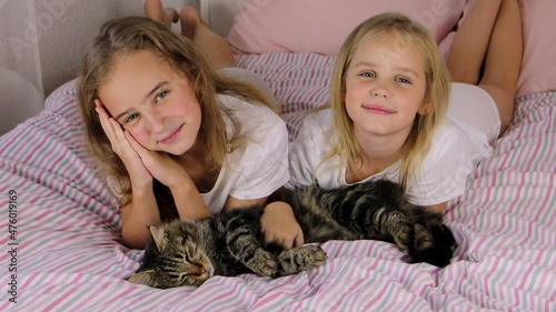 two cute european girls with blond hair are lying on the bed, looking at the camera and smiling, stroking a gray tabby domestic cat