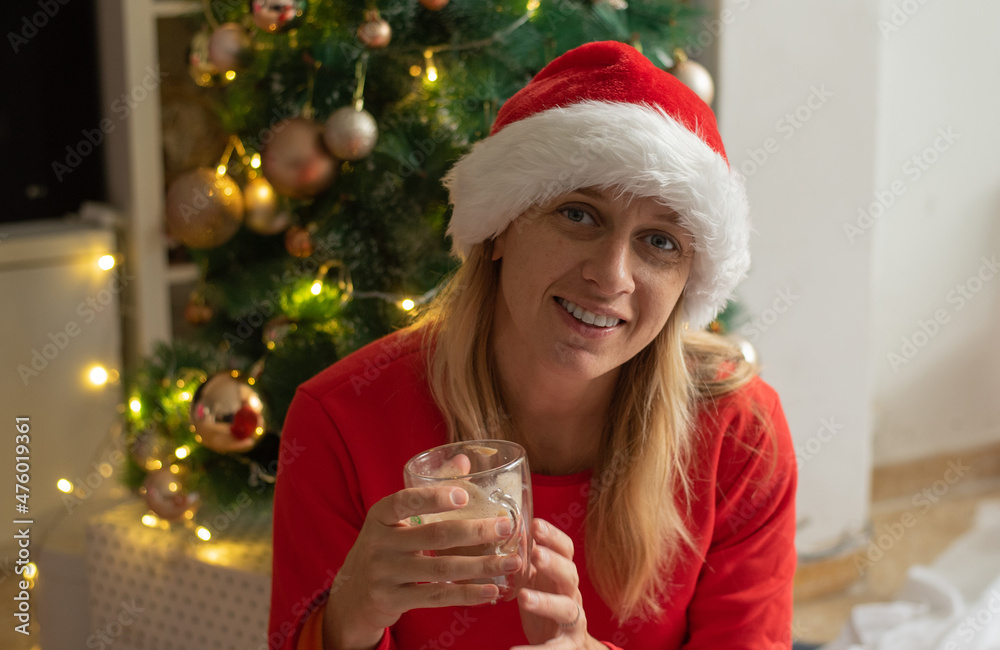 Woman wearing red pyjamas and Christmas hat. Santa girl with cup of coffee near Christmas tree. Feel fairy tale and magic. new year emotional and mood. Morning of holidays Woman wearing red pyjamas and Christmas hat. Santa girl with cup of coffee near Christmas tree. Feel fairy tale and magic. new year emotional and mood. Morning of holidays