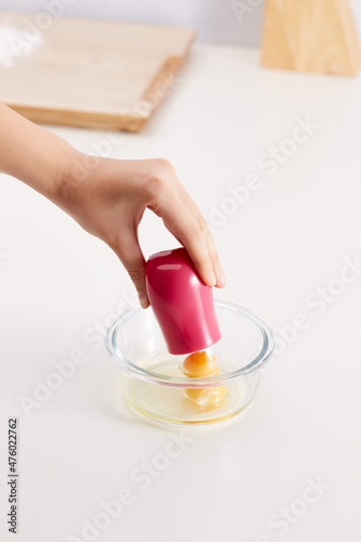 Shot of hand that holds a pink egg separator above transparent glass bowl. Bowl stands on the white table. A plate stands on the wooden board. Wooden items on the background. photo