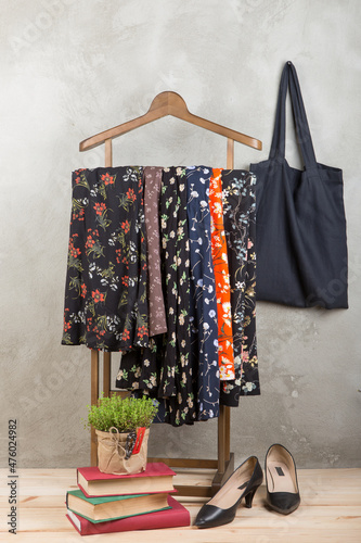 Shopping and style concept - clothes rack with trendy dresses in floral print, shoes and black eco tote bag on wooden floor and grey concrete background