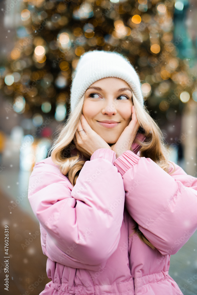 A cute blonde in a hat and a pink jacket against the background of a Christmas tree on the street. Winter walk, new year, feast day.