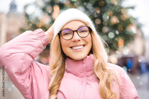 Cute portrait of a girl in glasses on the background of a Christmas tree. Winter, the first snow, a blonde in a pink jacket and a white hat smiles. New Year