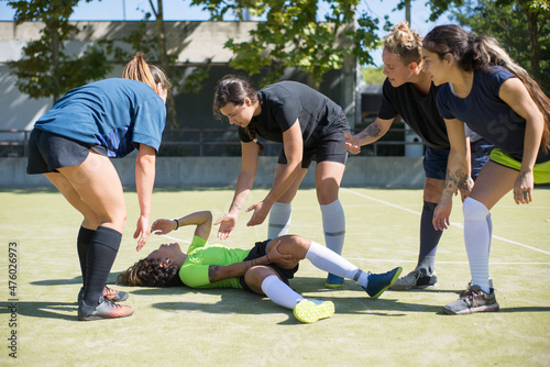 Woman with curly hair injured during football training. Sportswoman in sport clothes lying on ground, touching leg in pain, friends helping. Football, sport, leisure concept