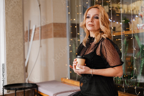 Young charming blonde with a cute smile and makeup while relaxing in a cafe. She is holding a cup of coffee in her hands. She is dressed in a black dress with transparent sleeves.