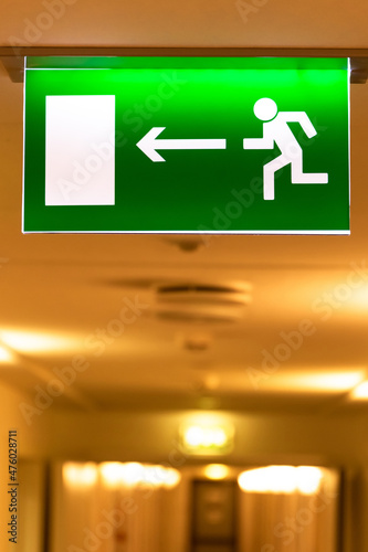 A bright green emergency evacuation sign with a directional arrow and a running figure hanging from the ceiling in the hallway. Selective focus. Close-up