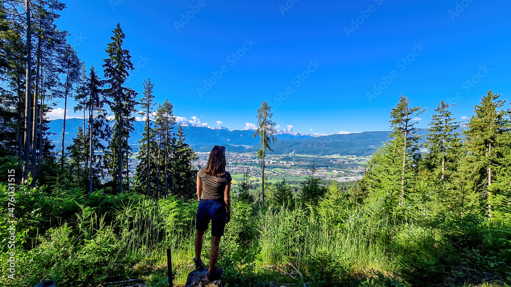 A woman standing in a forest in the Austrian Alps, Carinthia. The woman enjoys panoramic view on Villach and the Karawanks mountain chain in the back. Wanderlust, freedom and serenity.