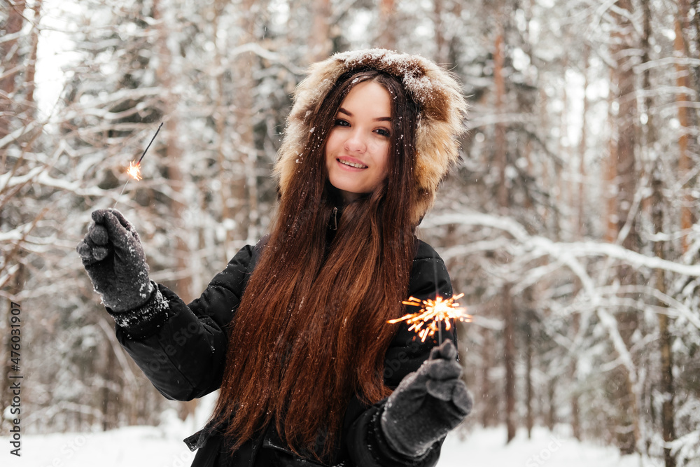 Woman in black coat holding sparkler in hand and smiling looking at camera. 20 years old girl in winter forest outdoors Woman in black coat holding sparkler in hand and smiling looking at camera. 20 years old girl in winter forest outdoors