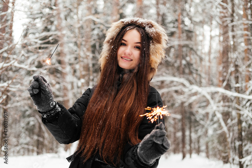 Woman in black coat holding sparkler in hand and smiling looking at camera. 20 years old girl in winter forest outdoors