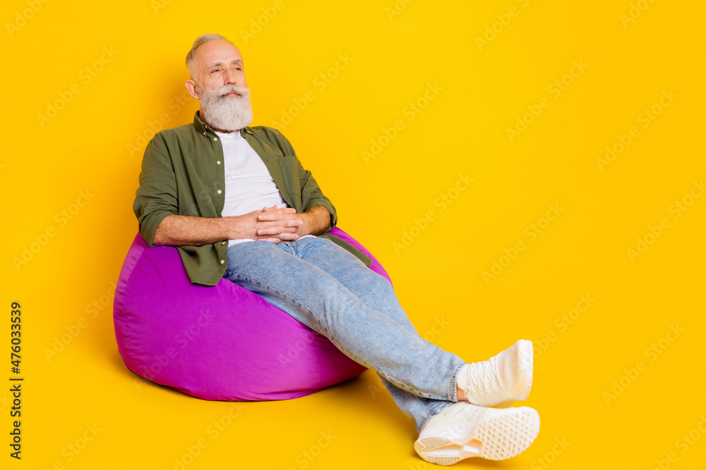 Photo of serious old man sit beanbag hold hands look empty space wear green shirt isolated yellow color background Photo of serious old man sit beanbag hold hands look empty space wear green shirt isolated yellow color background