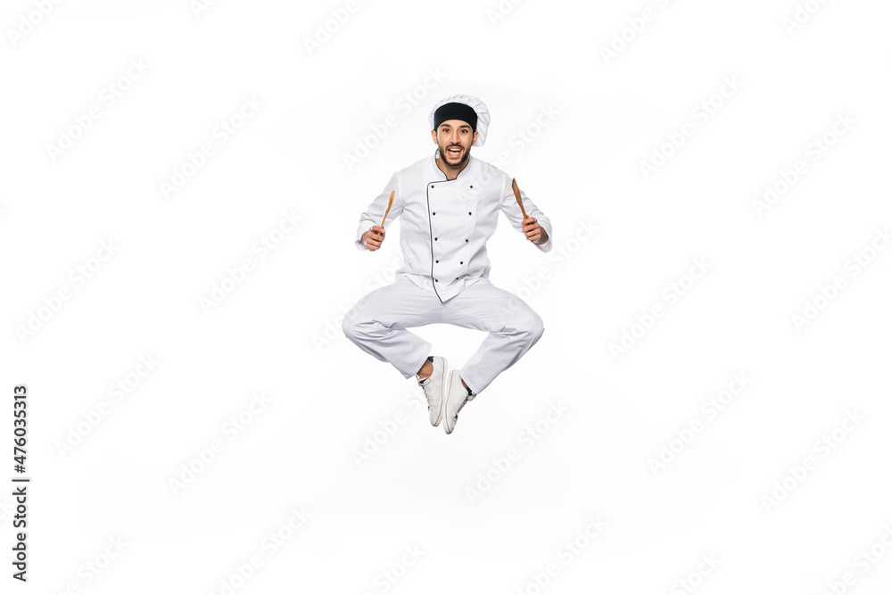 cheerful chef in hat and uniform jumping and holding wooden spatula and spoon isolated on white.