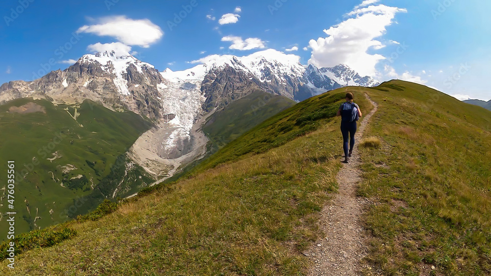 A woman on a hiking trail with a panoramic view on the snow-capped peaks of Tetnuldi,Gistola,Lakutsia and the Adishi Glacier in the Greater Caucasus Mountain Range in Georgia,Svaneti Region.Wanderlust