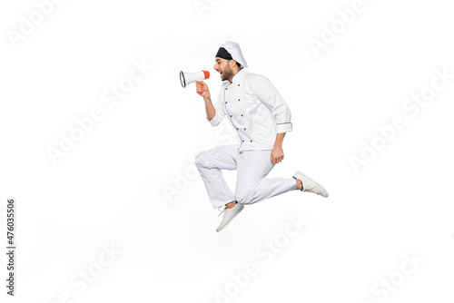 side view of young chef in hat and uniform levitating and speaking in megaphone isolated on white.