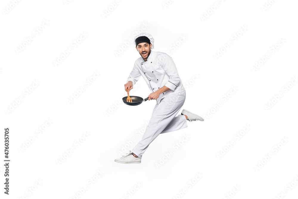 smiling chef in hat and uniform jumping and holding frying pan with wooden spatula isolated on white. smiling chef in hat and uniform jumping and holding frying pan with wooden spatula isolated on white.